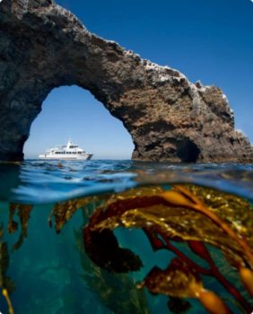 yate bajo un arco rocoso; la mitad de la imagen se encuentra bajo el agua cristalina, con algas en la parte inferior