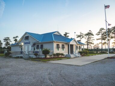 Small building with blue roof surrounded by trees and a gravel parking lot