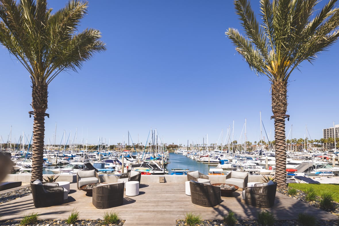 outdoor lounge chairs around two firepits with two large palm trees on a deck overlooking marina